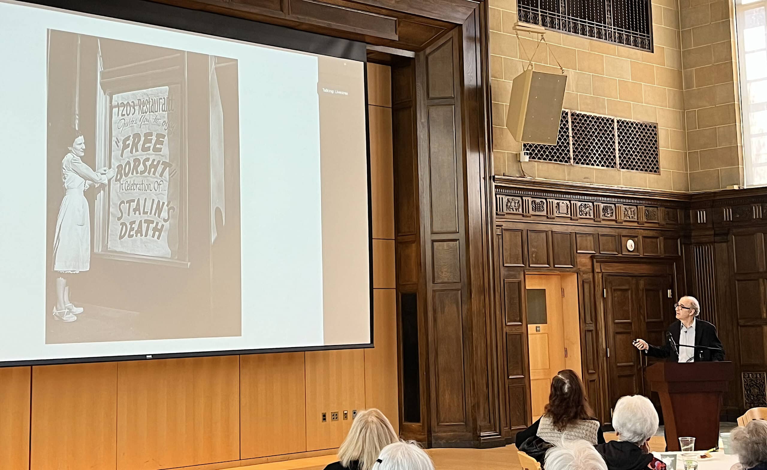 Joshua Rubenstein speking with slide of a restaurant window's sign Free Borscht to celebrate Stalin's death